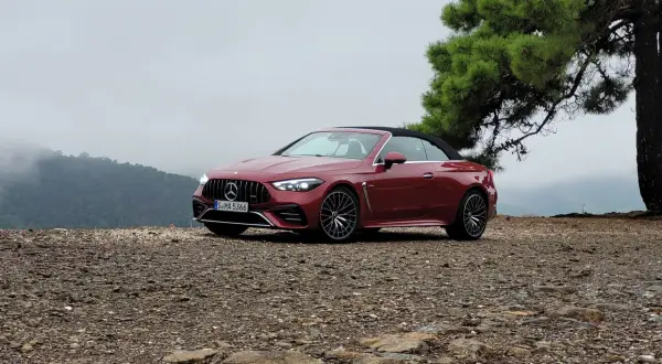 Czerwony kabriolet Mercedes-AMG CLE 53 na kamienistym podłożu, z drzewem i mglistymi górami w tle.