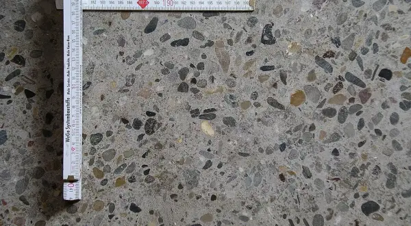 Superficie di calcestruzzo a vista con aggregati di varie dimensioni. Un righello pieghevole misura la **classe di esposizione calcestruzzo**.