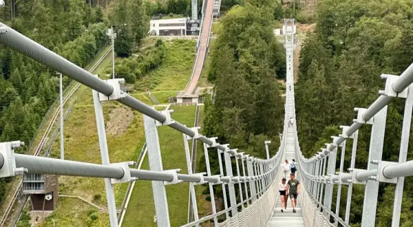 Menschen spazieren über den Willingen Skywalk, eine Hängebrücke mit Blick auf eine Skisprungschanze und bewaldete Hänge.