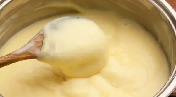 Une cuillère en bois prélève une généreuse portion de crème pâtissière onctueuse dans une casserole.