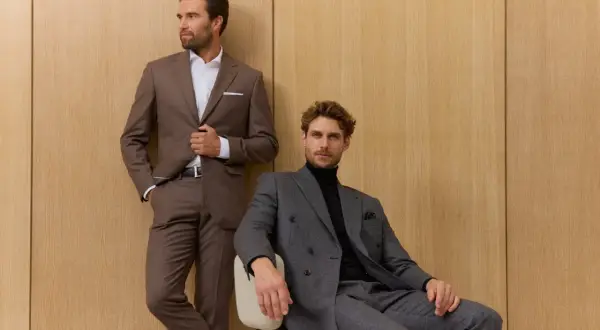Zwei Männer in eleganten Anzügen vor Holzvertäfelung. Der eine steht, der andere sitzt. Die perfekte sakko passform unterstreicht ihre maskuline Silhouette.