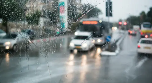 Gouttes de pluie sur un pare-brise, floutant la circulation urbaine. Les feux position seuls des voitures et des bus créent des traînées lumineuses sur la chaussée mouillée.