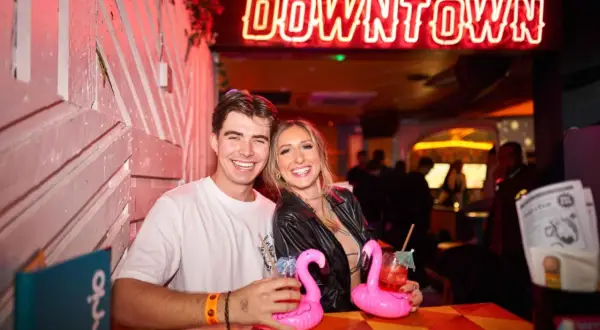 Coppia sorridente in un locale di Londra, con cocktail rosa e un'insegna al neon "DOWNTOWN".
