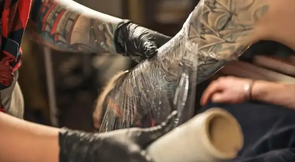 Manos enguantadas cubren un tatuaje recién hecho en el brazo con plástico. Aprende como cuidar un tatuaje recien hecho para una curación óptima.