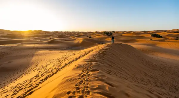 Ein einsamer Wanderer steht auf einer Düne während einer Marokko Sahara Tour. Fußspuren im Sand führen zum Horizont, wo die Sonne untergeht.