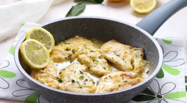 Scaloppine di pollo al limone in padella, guarnite con spicchi di limone fresco e prezzemolo tritato.