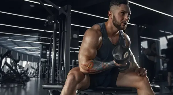 Muskularny mężczyzna z tatuażami ćwiczy biceps z hantlem w siłowni. Jego determinacja jest widoczna, jakby każdy trening był krokiem do celu, być może wspieranym przez suplementy takie jak clenbuterol.