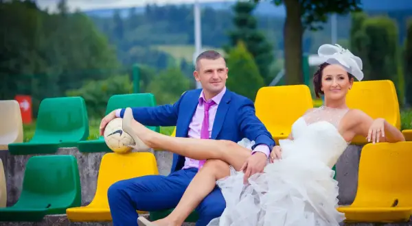Niespodzianka dla pary młodej: pan młody w garniturze i panna młoda w sukni ślubnej siedzą na trybunach, ona opiera nogę na piłce.