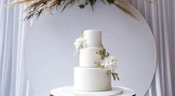 Eleganckie torty weselne wzory: biały, trzypiętrowy tort z delikatnymi kwiatami i koronkowym zdobieniem, prezentowany na złotym postumencie.