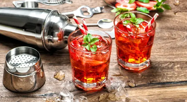 Zwei rote Cocktails mit Erdbeeren und Minze, Eiswürfel, Cocktailshaker und Barzubehör auf Holztisch.