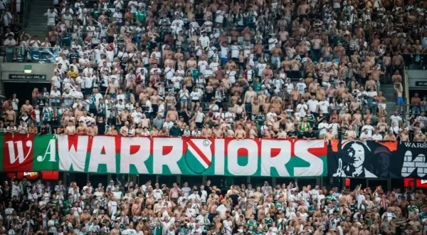 Tłum kibiców Legii Warszawa, z transparentem "WARRIORS" i herbem klubu. Wszyscy w biało-zielonych barwach, gotowi do dopingu.