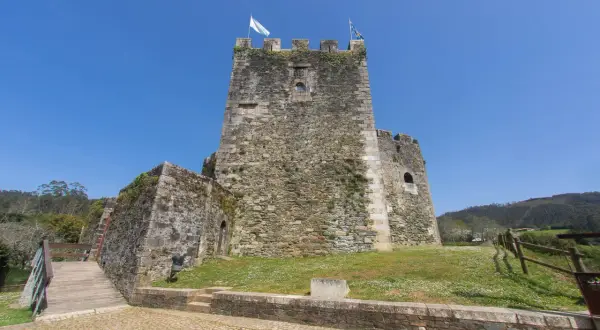 El castillo de Moeche, una imponente fortaleza de piedra, se alza bajo un cielo azul. Un puente de madera conduce a sus muros cubiertos de vegetación.
