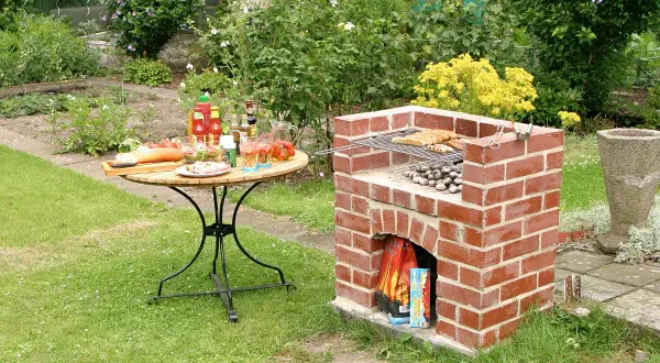 Ein selbstgebauter Ziegelgrill mit Würstchen und Kartoffeln, bereit für ein Gartenfest. Ein Tisch mit Salaten und Getränken steht daneben.