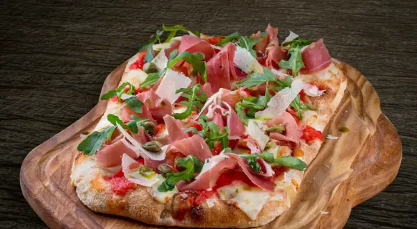 En avlång pinsa pizza med prosciutto, ruccola och parmesan på en träbricka.