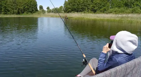 Osoba siedzi nad jeziorem Zagnanie, łowiąc ryby. W tle zielone drzewa i trzciny.