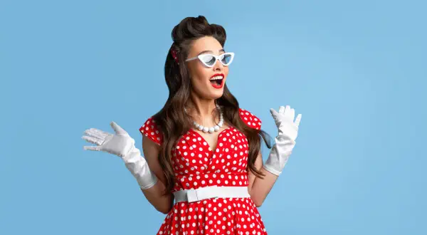 Kobieta w czerwonej sukience w grochy, białych rękawiczkach i okularach w stylu pin up girl stylizacje, z uniesionymi rękami.