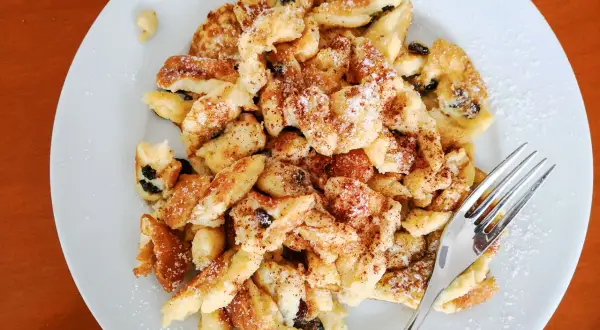 Ein Teller mit fluffigem Kaiserschmarrn mit Eischnee, bestreut mit Puderzucker und Zimt. Daneben eine Gabel.