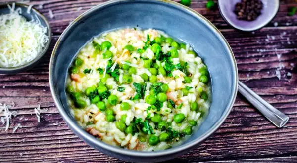 Ein cremiges Risi Bisi Rezept original, garniert mit frischer Petersilie und Erbsen. Daneben eine Schale geriebener Parmesan.
