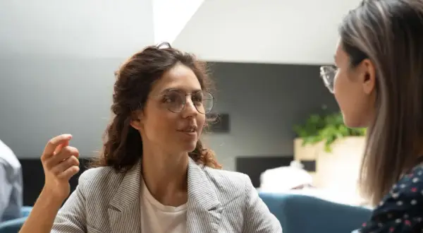 Två kvinnor i samtal, en med glasögon och lockigt hår, den andra med långt hår. En man syns i bakgrunden. Scenen utstrålar autentiskt ledarskap genom öppen kommunikation.