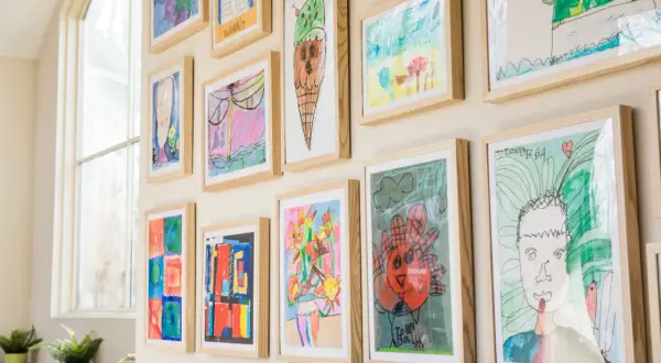 A vibrant kids art gallery wall showcases colorful drawings of ice cream, flowers, and portraits, each framed in light wood.