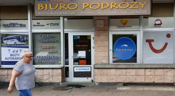 Biuro podróży z ofertą Grecos. Kobieta w paski idzie chodnikiem przed witryną z logo Grecos.