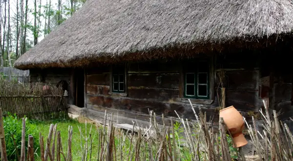 Drewniana chata ze słomianym dachem, otoczona płotem z gałęzi i glinianym dzbanem.