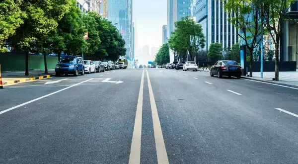 Eine Straße mit einer **durchgezogenen Linie** in der Mitte, gesäumt von Bäumen und modernen Gebäuden. Autos parken am Straßenrand.