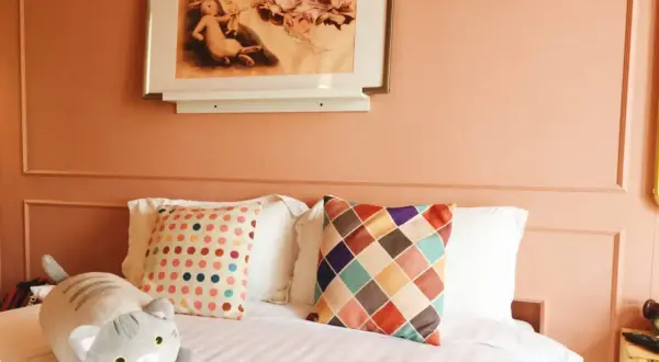 Chambre couleur terracotta avec un lit douillet, des coussins colorés et une peluche chat. Un tableau de lapins orne le mur.