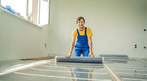 Uśmiechnięty pracownik w niebieskim ogrodniczkach rozkłada folię grzewczą na podłodze.