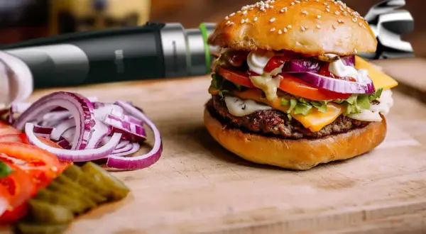 Pyszny hamburger z serem, warzywami i domowym sosem do hamburgerów. Obok pokrojona cebula i ogórki.