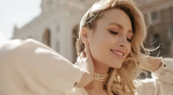 Uśmiechnięta blondynka z perłową opaską i naszyjnikiem. Różne rozmiary łańcuszków podkreślają jej szyję.