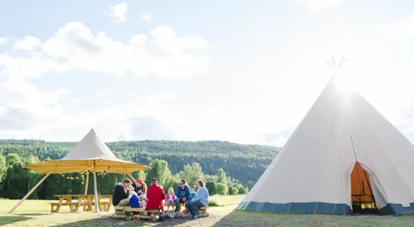 En familj njuter av en solig dag på alevi camping i Sverige. De sitter runt en lägereld under ett stort parasoll, med en tipi i bakgrunden.