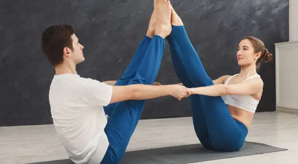 Pareja practicando yoga juntos, realizando posturas para hacer con tu pareja. Se miran sonriendo mientras unen sus pies.