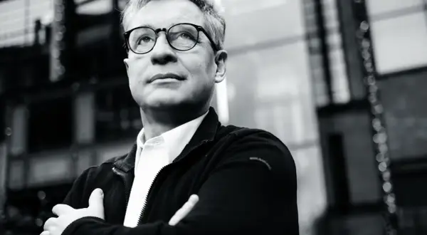Mężczyzna w okularach, z rękami skrzyżowanymi na piersi, patrzy w górę. Jego postawa sugeruje, że jest alior ostrożny, analizując sytuację.