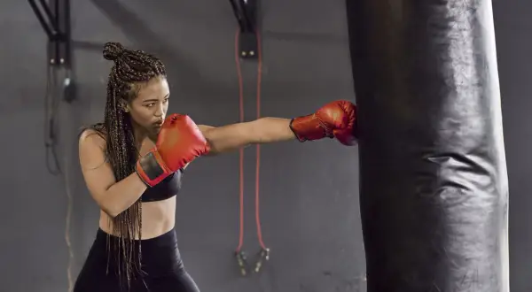 A woman with braided hair throws a punch at a heavy bag, proving that boxing tones arms and builds strength.