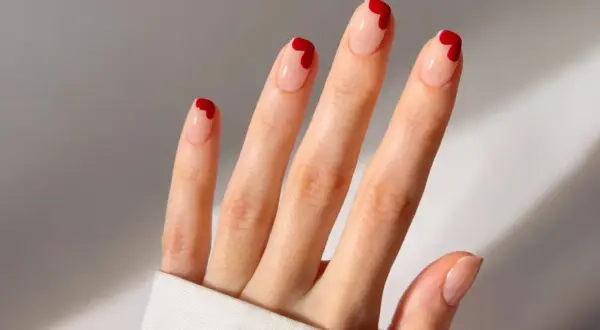 Une main aux ongles nude ornés de petits cœurs rouges. Une idée ongle coeur parfaite pour la Saint-Valentin ou pour ajouter une touche d'amour à votre quotidien.