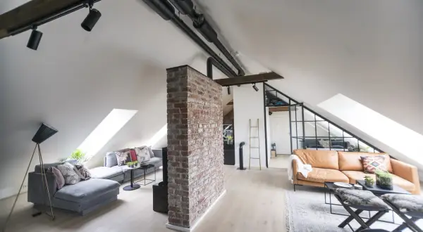 Un salon mansardé avec une cloison de séparation sous pente en briques apparentes, deux canapés, des luminaires industriels et des fenêtres de toit.