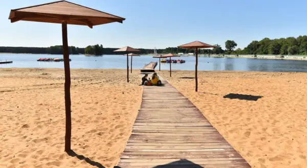 Sielpia jezioro: drewniana ścieżka prowadzi na piaszczystą plażę pod parasolami. W oddali widać pomost z rowerami wodnymi.
