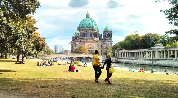 Ett par promenerar på en gräsmatta med Berlin domkyrka i bakgrunden. Många njuter av solskenet, en perfekt dag för aktiviteter i Berlin.
