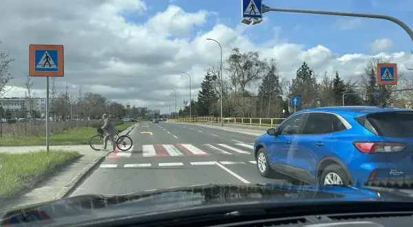Niebezpieczne wyprzedzanie na pasach na drodze dwupasmowej. Niebieski SUV omija rowerzystkę stojącą przy przejściu.