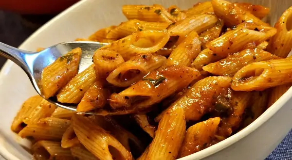 Eine Löffelportion Penne all'Arrabiata, mit einer würzigen Tomatensauce und Kräutern, wird aus einer Schüssel gehoben.