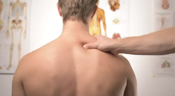 Dłoń terapeuty masuje bark mężczyzny na tle plakatów anatomicznych.