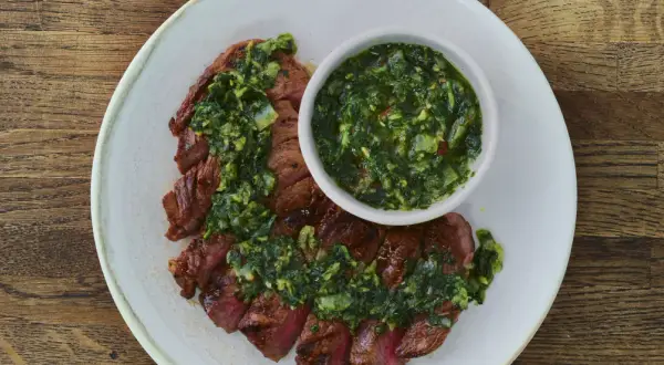 Pokrojony stek z wołowiny polany zielonym sosem chimichurri, podany na talerzu.