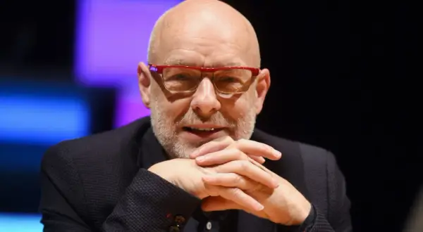 Brian Eno, eno musicista inglese, con occhiali rossi e mani intrecciate.