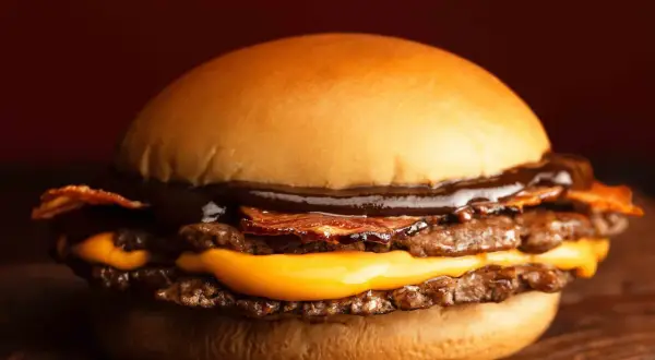 Gros plan sur un burger double viande, fromage fondant, bacon croustillant et sauce barbecue, sur une planche en bois.