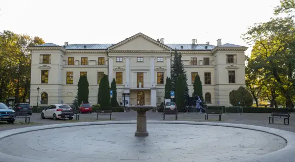 Pałac Dernalowiczów z fontanną i ławkami przed fasadą. W tle drzewa i zaparkowane samochody.