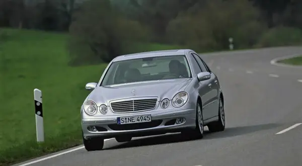 Srebrny Mercedes W211 z 2004 roku jedzie po krętej drodze wśród zielonych wzgórz.