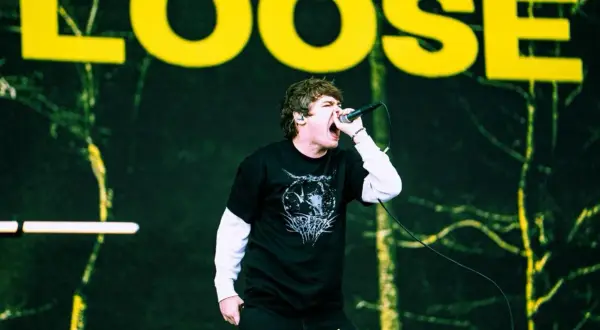 Wokalista z otwartymi ustami śpiewa do mikrofonu na tle sceny z napisem "LOOSE".