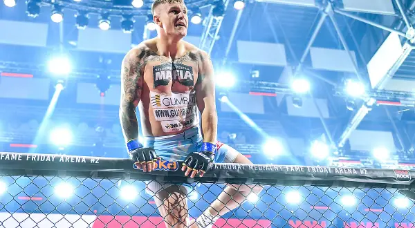 Alan Kwieciński, wytatuowany zawodnik MMA, w rękawicach, oparty o siatkę klatki, w świetle reflektorów.