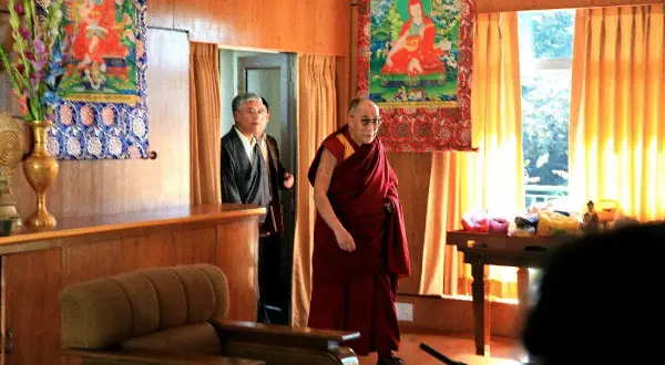 Dalai Lama i sin klädsel, med en man i kostym bakom. En inblick i var bor Dalai Lama, med buddhistisk konst på väggarna.